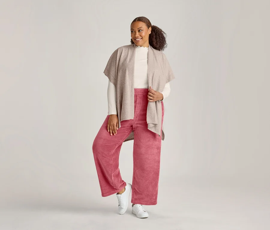 Une femme souriante se tient debout, vêtue d'un poncho beige, d'un haut clair, d'un pantalon rose large et de baskets blanches.