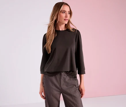 Une femme aux longs cheveux blonds porte un t-shirt en jersey 3/4 Street One marron et un pantalon à motifs devant un fond rose.