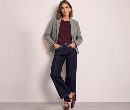 Une femme debout les mains dans les poches, portant un blazer Street One à carreaux gris sur un haut bordeaux, un jean large bleu foncé et des chaussures plates rouge foncé, appuyée contre un mur clair.
