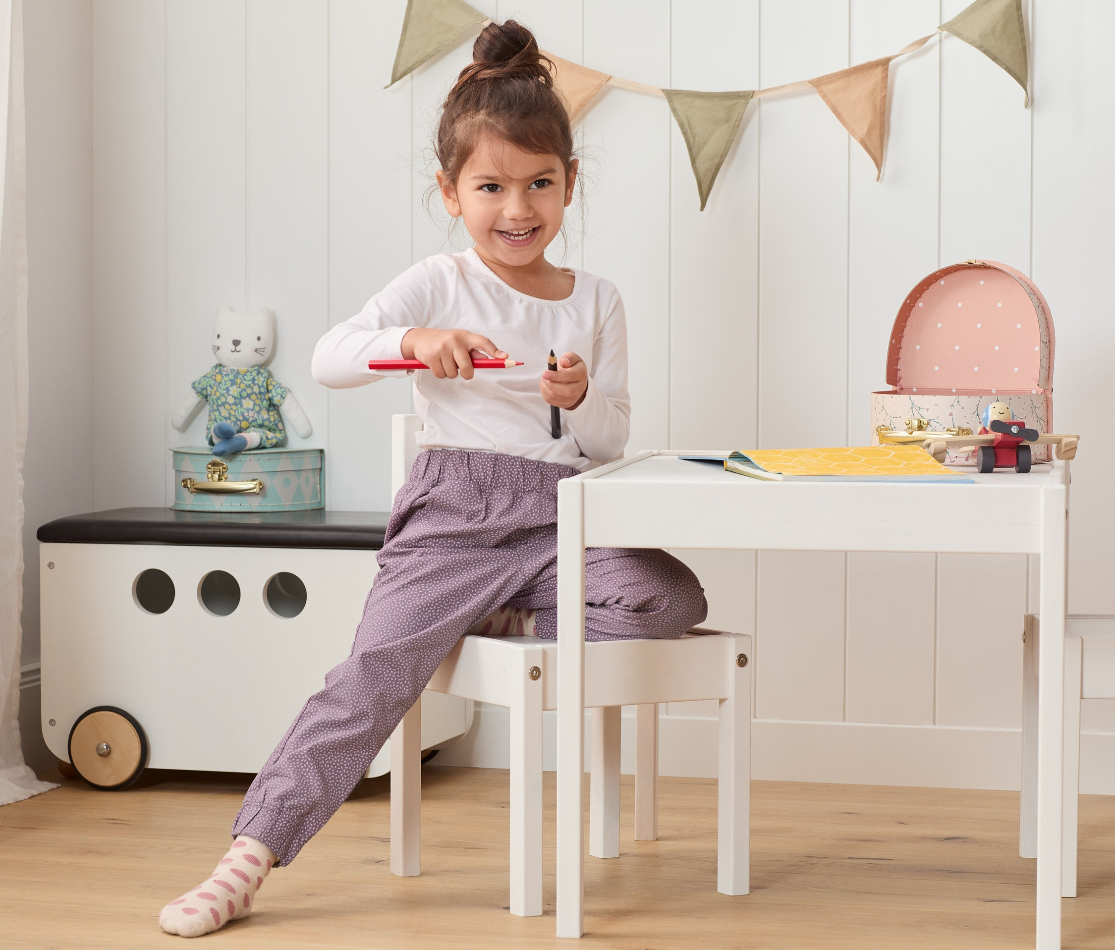 Fillette assise à une table tenant des crayons. Elle porte un pantalon à enfiler en coton.