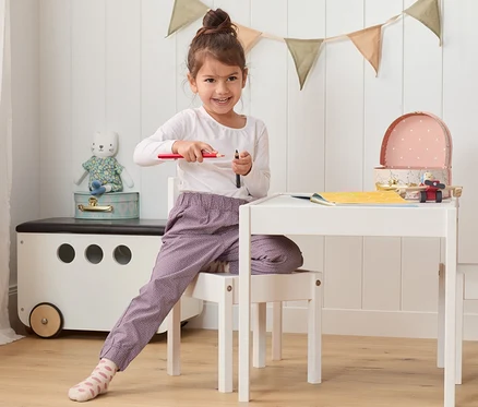 Fillette assise à une table tenant des crayons. Elle porte un pantalon à enfiler en coton.