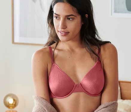 Une femme porte un soutien-gorge à armatures bois de rose et un cardigan dans une chambre.