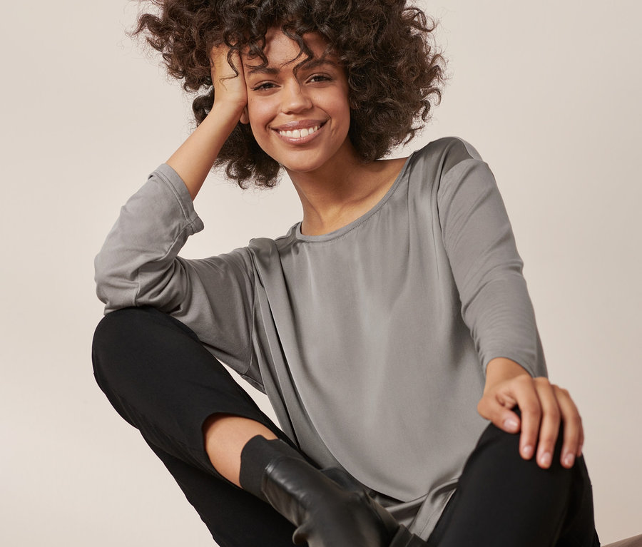 Femme assise posant en blouse bimatière, pantalon noir et bottes.