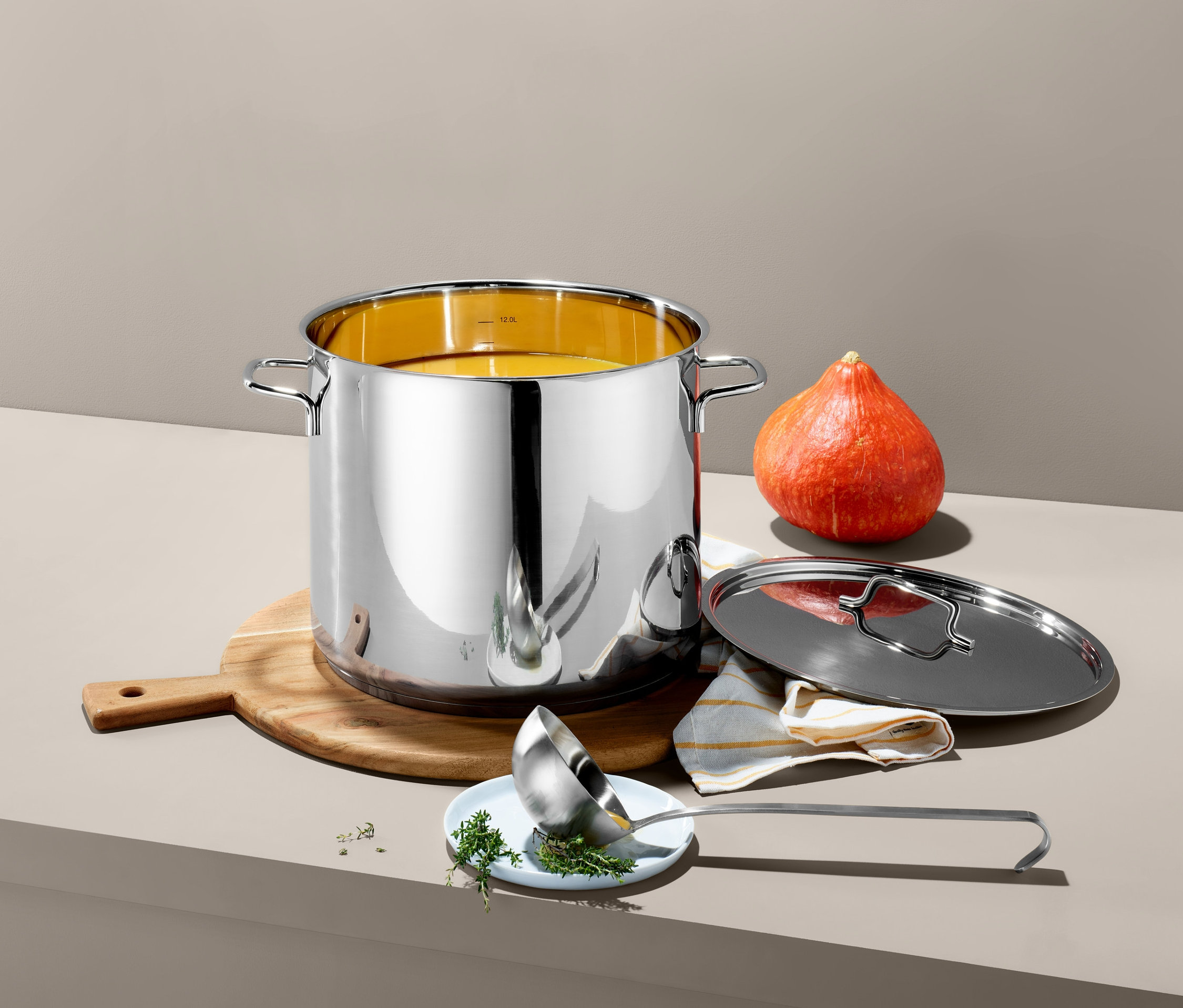 Une marmite en acier inoxydable XL est posée sur une planche à découper en bois à côté d'une courge. Un couvercle et une louche sont posés à côté de la marmite.