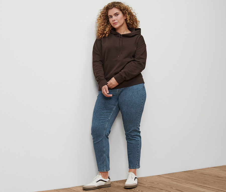 Une femme debout porte un jean droit en denim « Fit Juna » bleu moyen, des baskets en cuir et un sweat-shirt à capuche marron foncé.