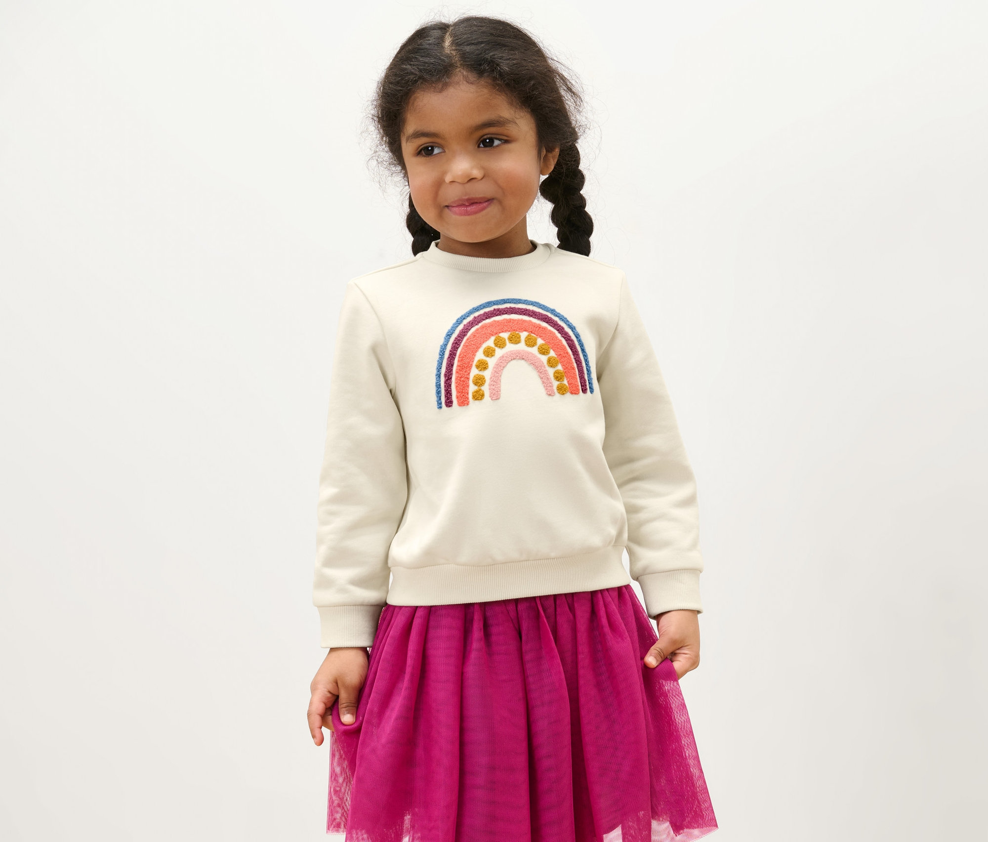 Fillette portant un sweat-shirt pour enfant avec motif arc-en-ciel appliqué et une jupe violette.