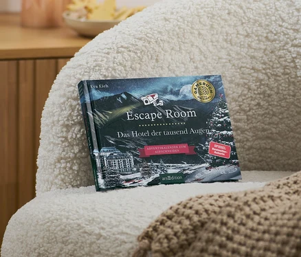 Sur un fauteuil se trouvent un livre calendrier de l’Avent Escape Room « Das Hotel der tausend Augen » (L’hôtel aux milles yeux, en allemand) et une couverture beige.