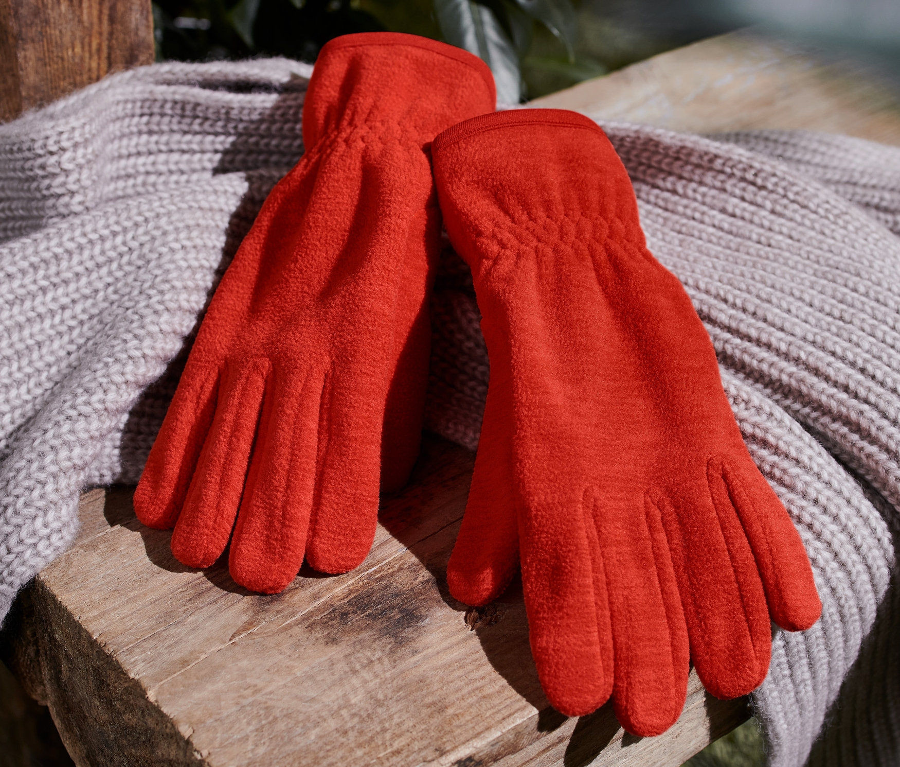 Des gants en polaire orange reposent sur un châle en tricot sur un banc en bois.