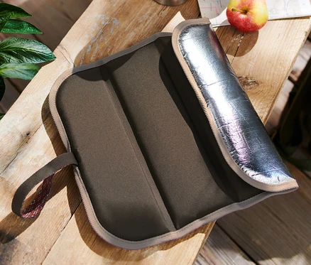 Un coussin d’assise thermique pliable est posé sur une table en bois à côté d’une gourde, d’une pomme et d’un sac à dos.