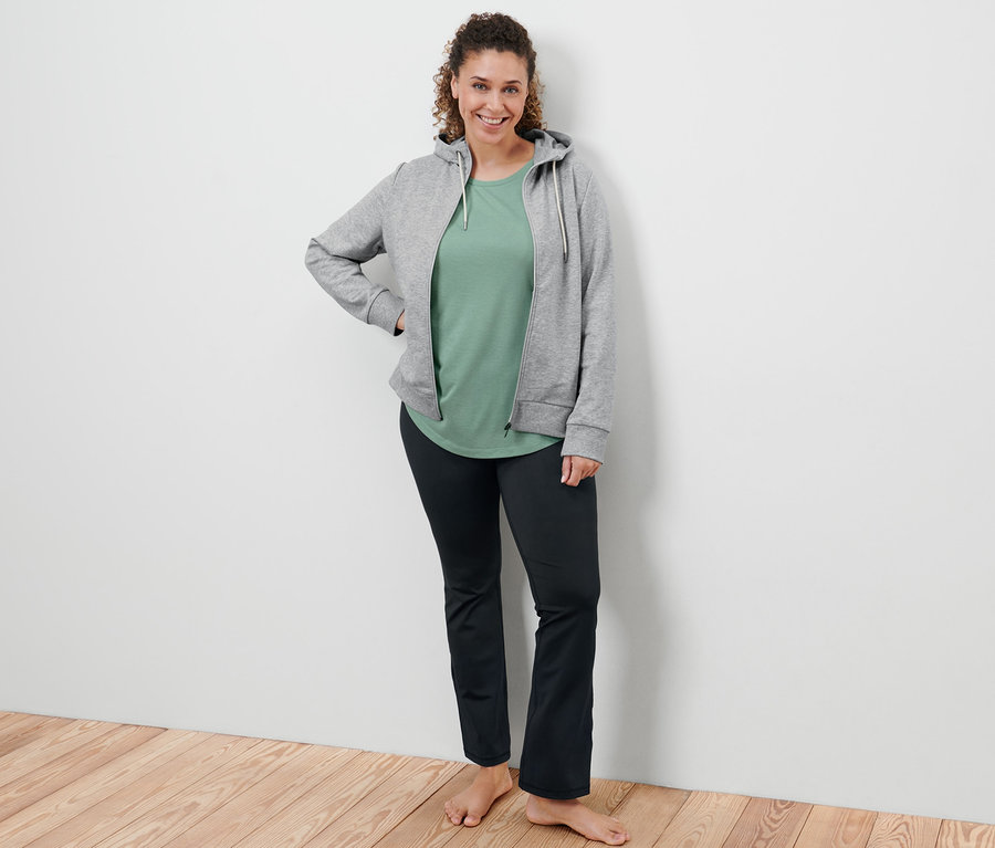Femme posant en veste molletonnée grise, t-shirt long vert sauge et pantalon de sport modelant.