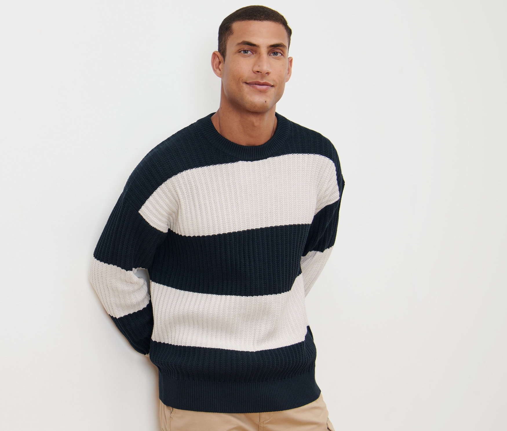 Un homme pose sur un fond blanc portant un pull en grosse maille rayé marine et blanc avec un pantalon kaki.