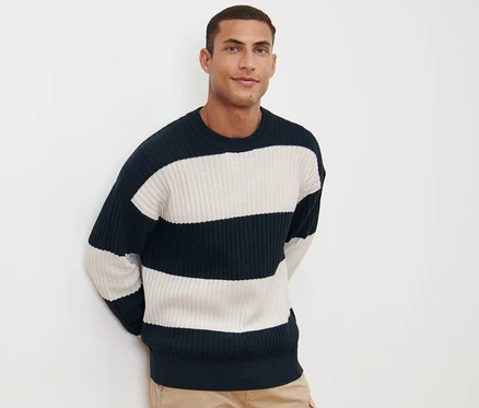 Un homme pose sur un fond blanc portant un pull en grosse maille rayé marine et blanc avec un pantalon kaki.