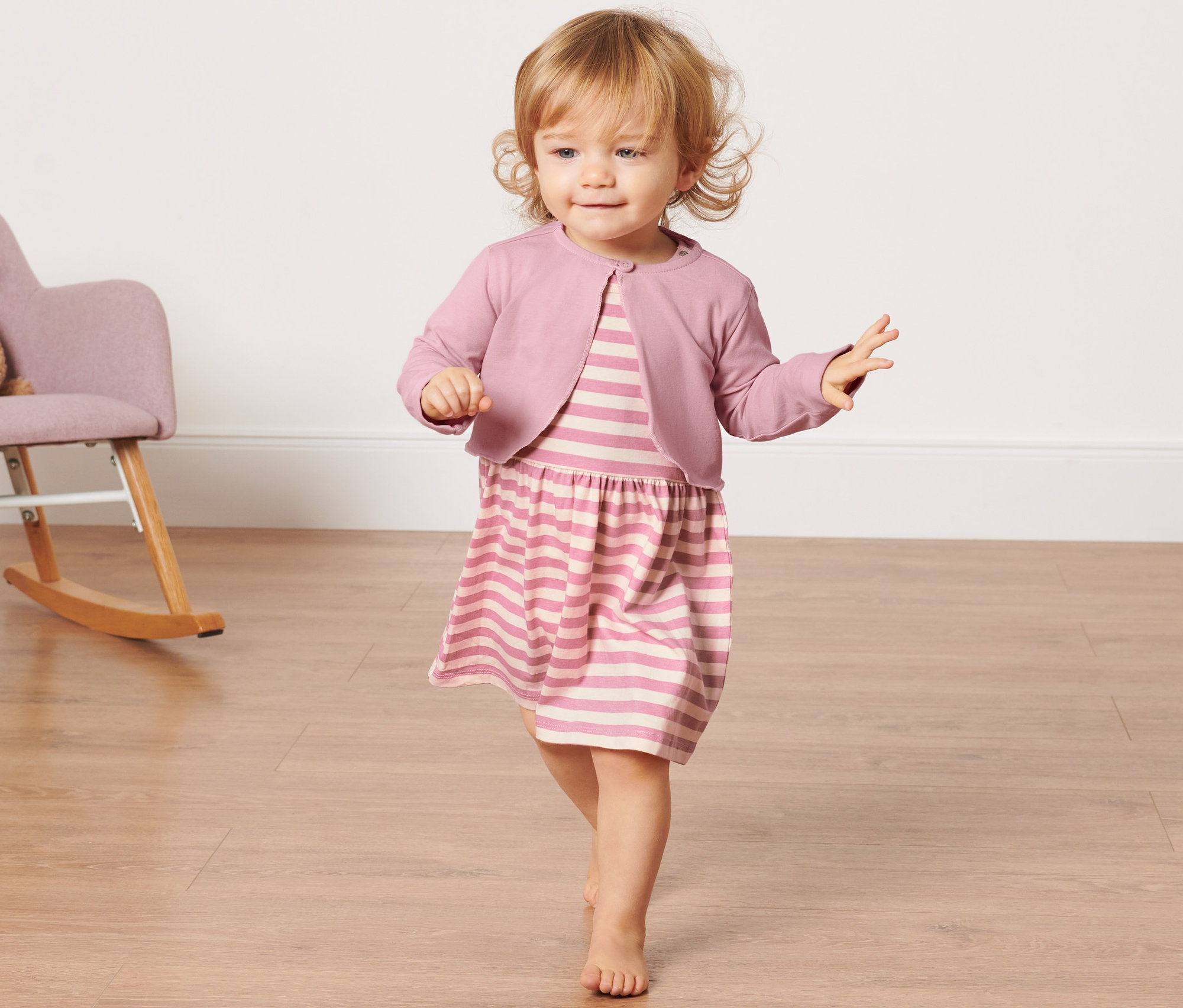 Un bébé en robe rayée et veste boléro pour bébé marche sur un plancher en bois. À l'arrière-plan, il y a un fauteuil à bascule avec un ours en peluche.