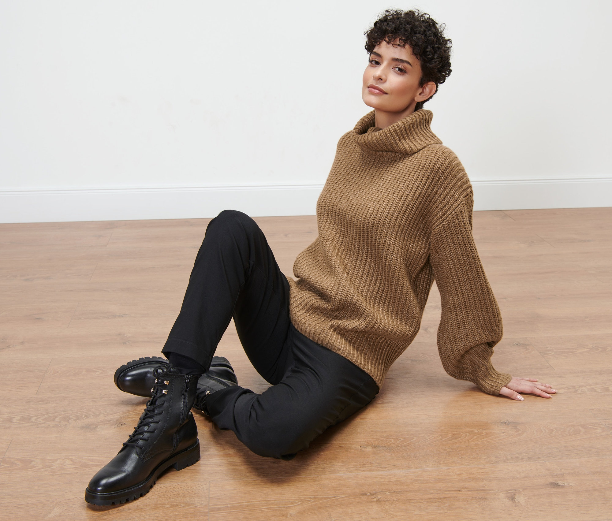 Femme assise par terre portant un pull à col roulé en grosse maille marron, un pantalon noir et des bottes noires.