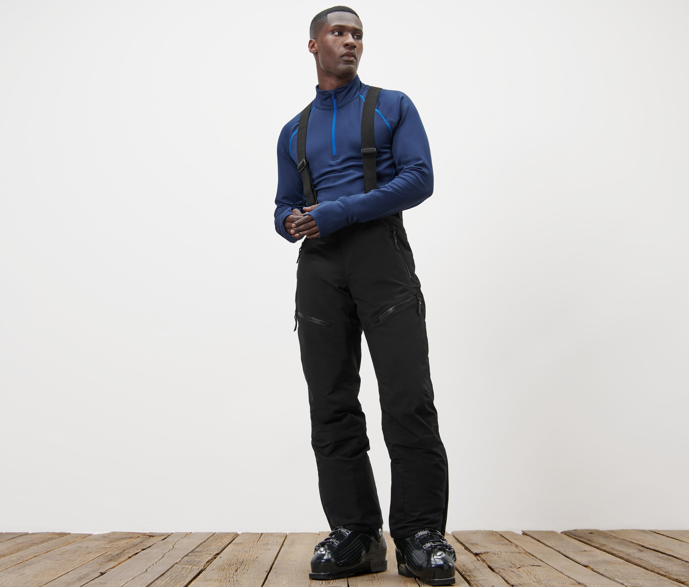 Un homme debout sur un plancher en bois porte un haut bleu et un pantalon de ski high-tech.