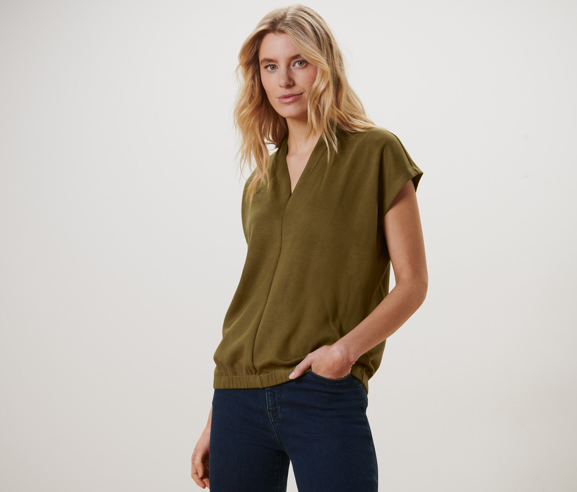 Blonde portant un chemisier olive à col en V et un jean bleu marine, posant.