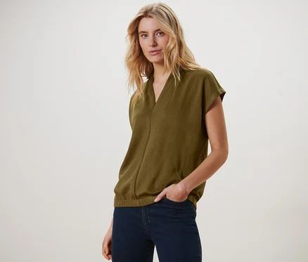 Blonde portant un chemisier olive à col en V et un jean bleu marine, posant.