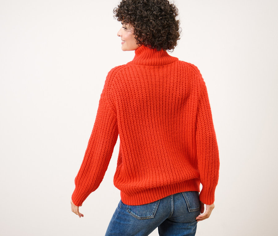 Vue arrière d'une femme aux cheveux bouclés portant un pull camionneur orange en grosse maille et un jean bleu.