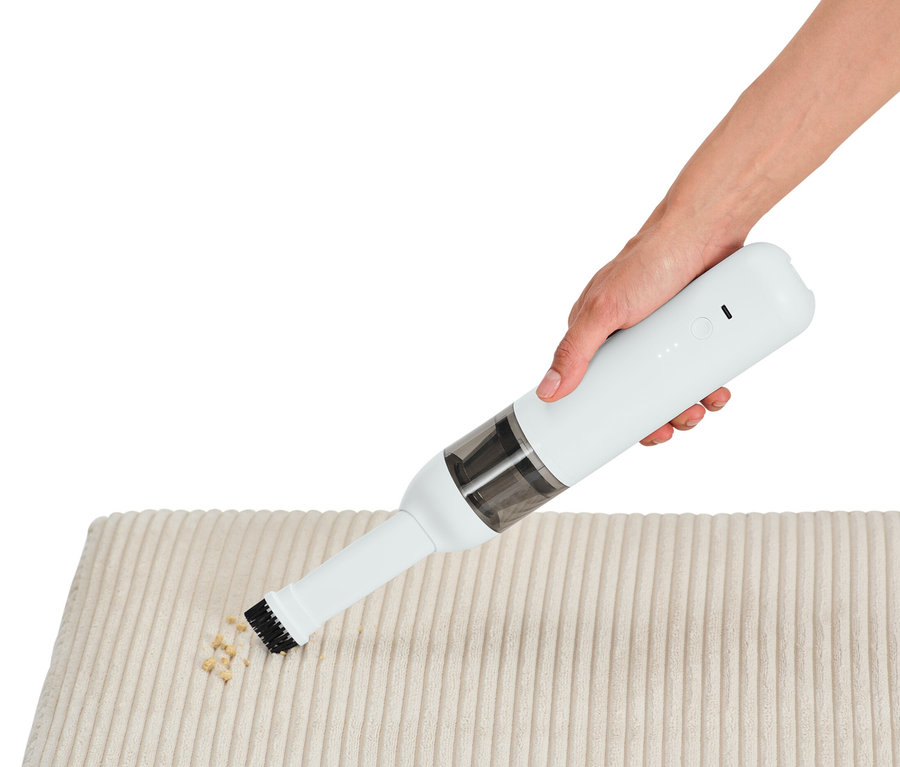 Une main utilise un aspirateur à main à batterie pour nettoyer des miettes sur un tissu crème.