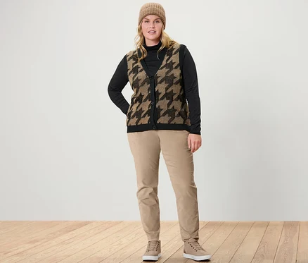 Une femme debout porte un t-shirt col roulé noir, une veste sans manches en maille à jacquard pied-de-poule et un pantalon en velours camel.