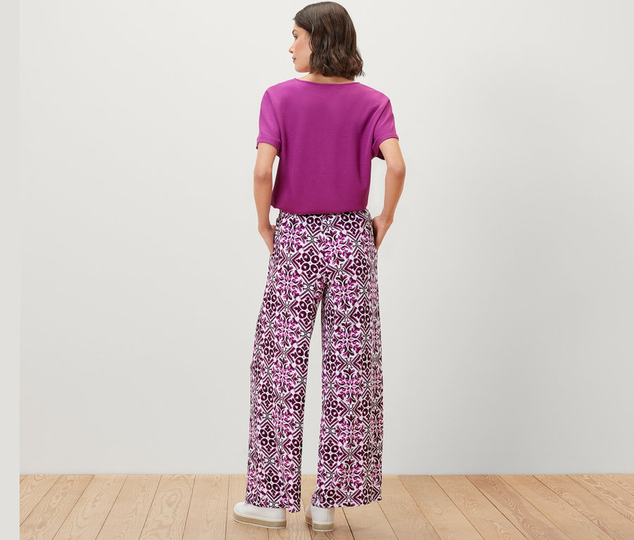 Une femme porte un pantalon palazzo imprimé de couleur baie et un t-shirt violet.