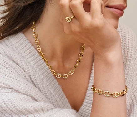 Une femme les yeux fermés touche son oreille, elle porte des créoles ovales avec des cristaux de verre, une bague «
A
cre
A
» avec des cristaux de verre, un collier «
A
cre
A
» avec des cristaux de verre et un bracelet «
A
cre
A
» avec des cristaux de verre.