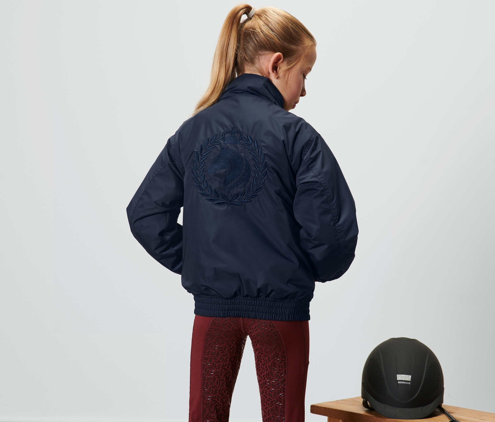 Une fille vue de dos porte un blouson pour enfant bleu foncé avec une broderie de cheval. Un casque d'équitation est posé sur un banc à côté d'elle.