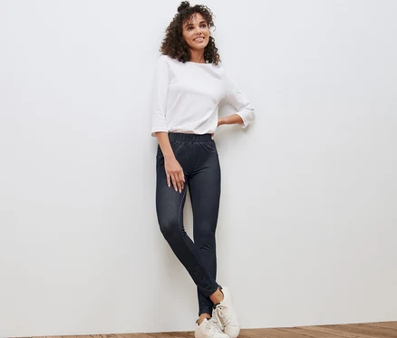 Une femme appuyée contre un mur blanc porte un legging aspect jean, un chemisier blanc et des baskets.