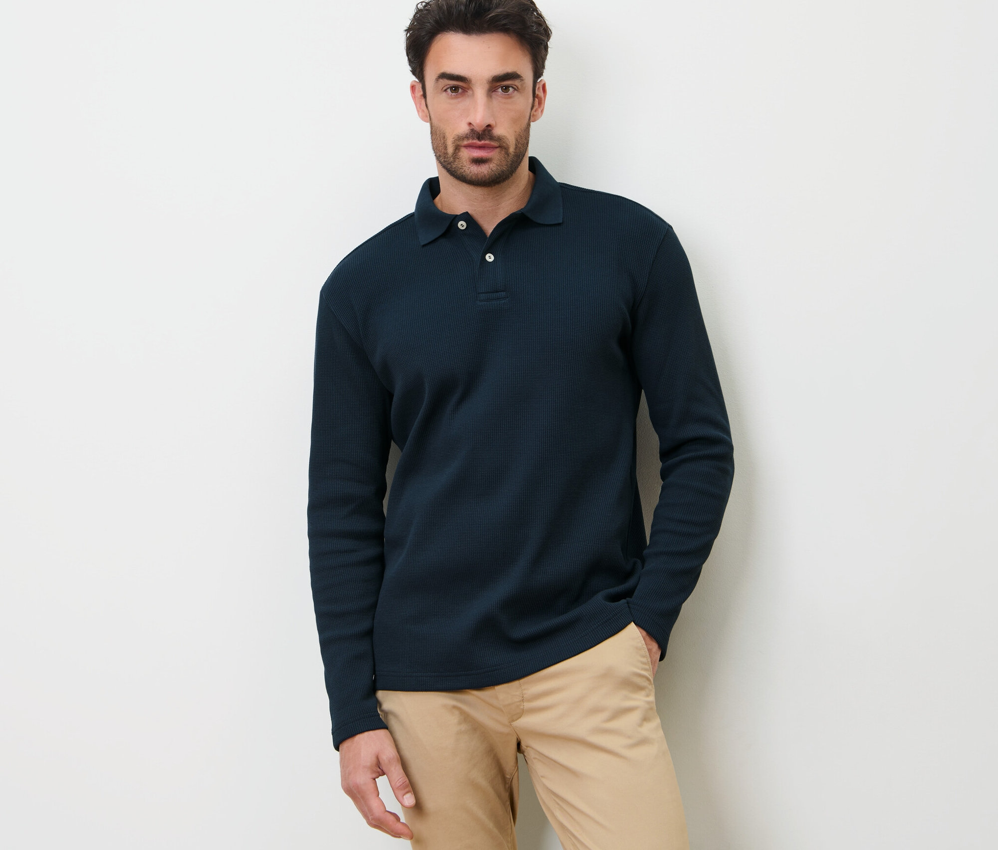 Un homme pose devant un fond blanc en polo manches longues bleu marine.