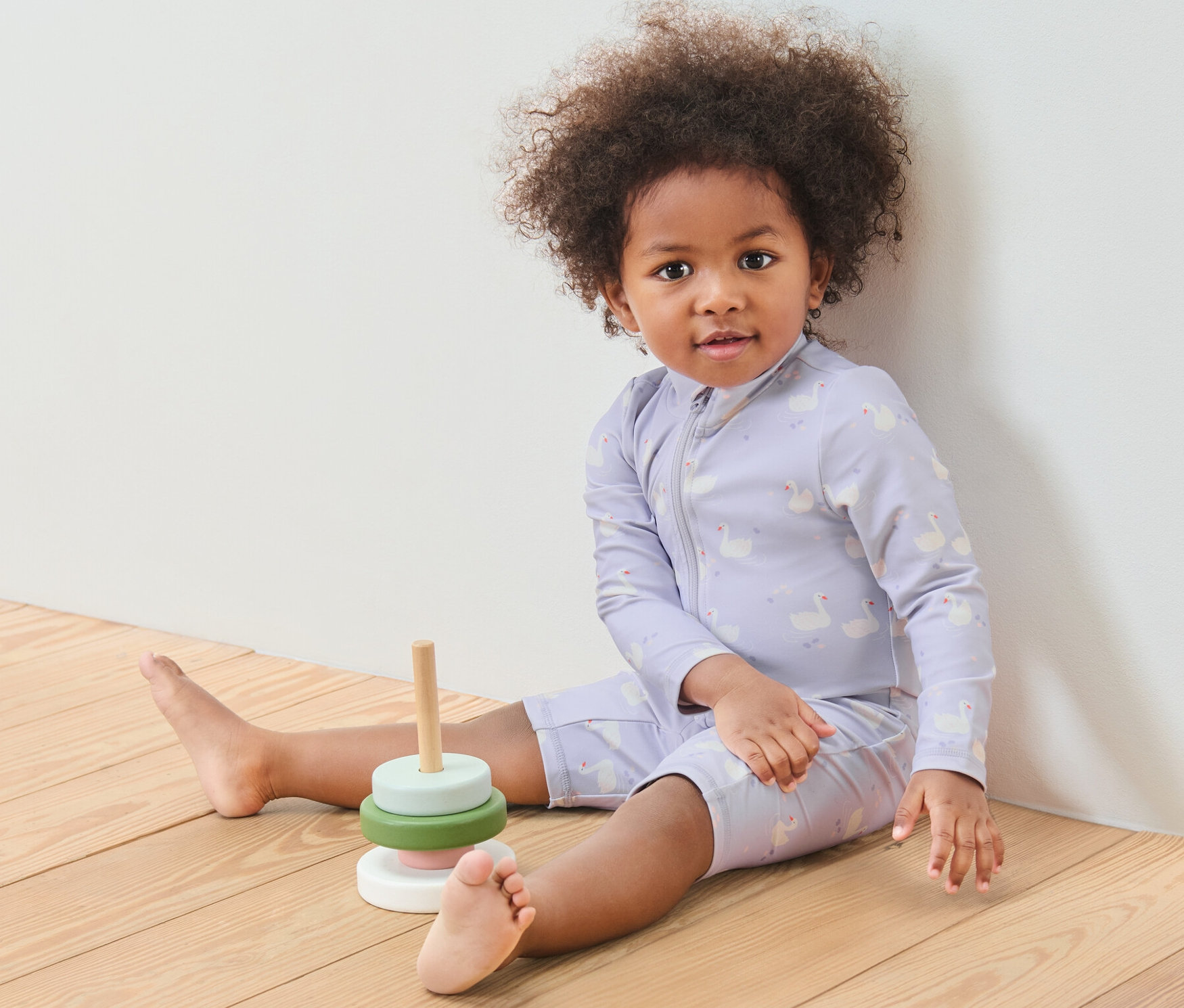 Un enfant est assis sur un plancher en bois à côté d'un jouet en bois. Il porte une combinaison pour bébé avec un indice de protection UV 80, cygne.