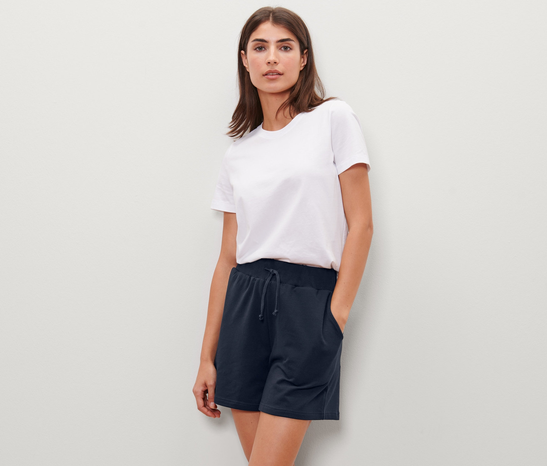 Une femme pose, portant un short molletonné bleu marine et un t-shirt blanc.