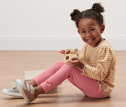 Une petite fille souriante est assise par terre et joue avec un appareil photo en bois. Elle porte un t-shirt rayé et 5 leggings pour enfant motif intégral.