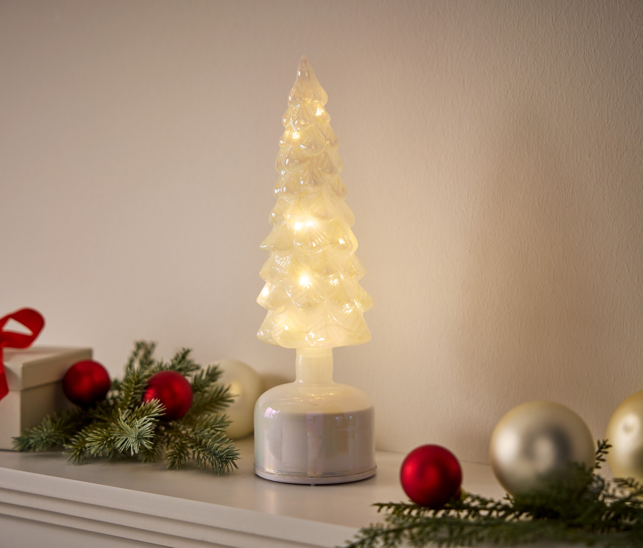 Un arbre décoratif illuminé est placé à côté d'un cadeau et de décorations sur une surface blanche.