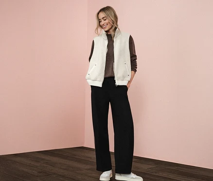 Une femme porte une veste en teddy Street One sur un pull marron, associé à un pantalon noir et des baskets blanches.