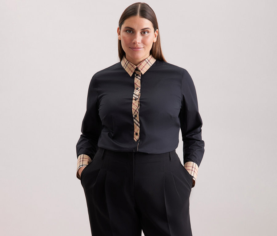 Une femme aux cheveux bruns porte une chemise en popeline à carreaux Seidensticker noire et un pantalon noir.