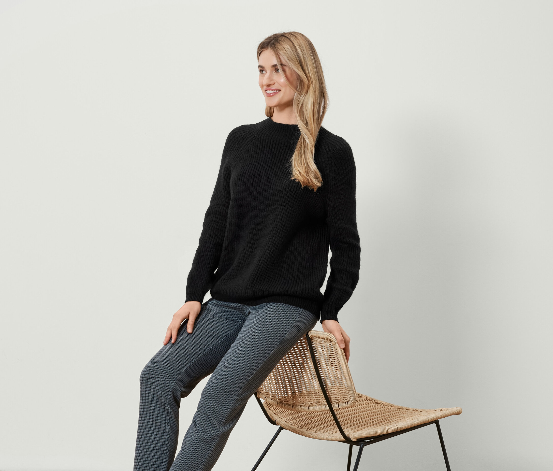 Une femme blonde est assise de côté sur une chaise en osier, portant un pull noir et un pantalon à motifs.