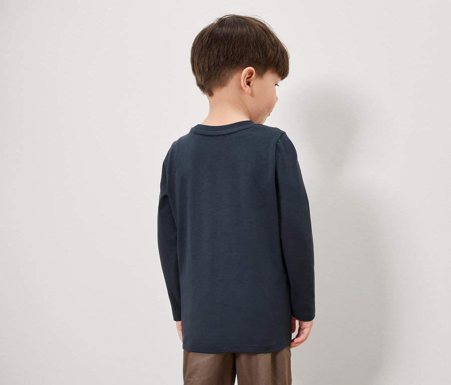 Un garçon, dos à la caméra, porte un t-shirt à manches longues bleu marine et un pantalon marron.