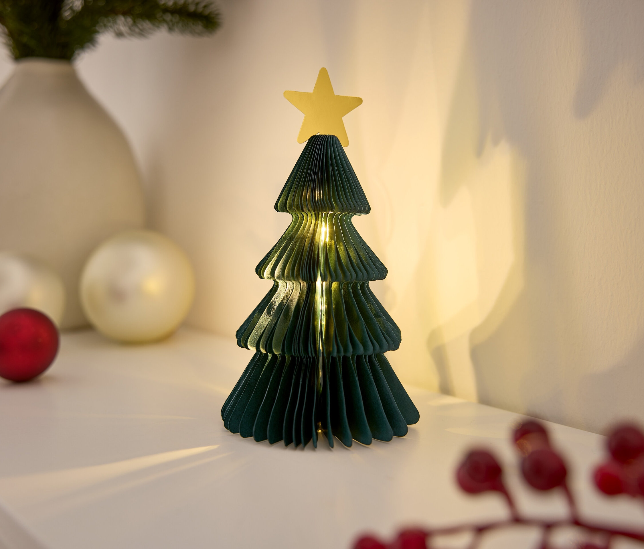 Sapin de Noël décoratif en papier avec une étoile sur une table blanche, illuminé par des lumières.