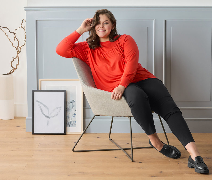 Une femme est assise sur une chaise et porte un pantalon stretch à carreaux, des loafers en cuir et une blouse avec fronces, orange-rouge.
