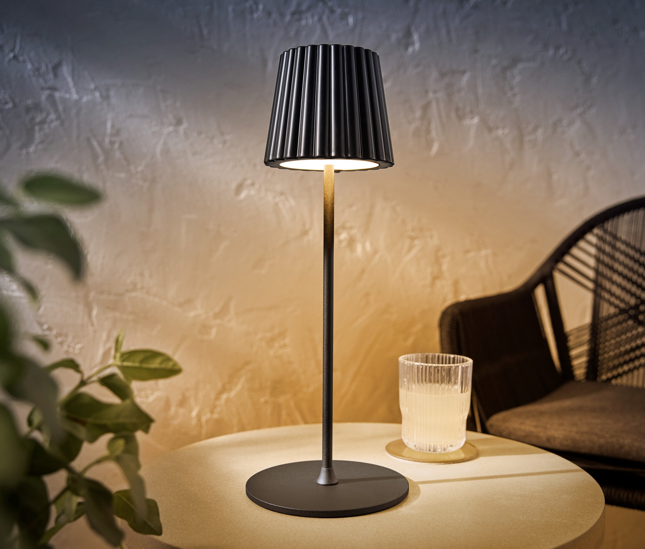 Une lampe de table d’extérieur à LED rechargeable noire est posée sur une table à côté d’un verre avec une boisson.