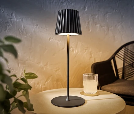 Une lampe de table d’extérieur à LED rechargeable noire est posée sur une table à côté d’un verre avec une boisson.