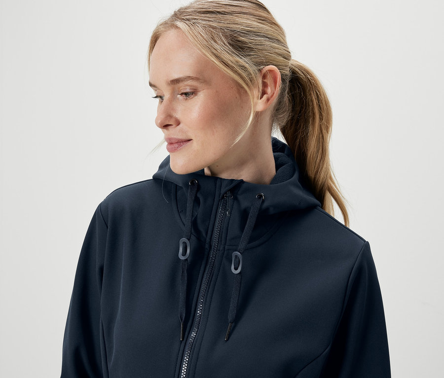 Portrait d'une femme blonde aux cheveux attachés en queue de cheval, portant un sweat à capuche bleu marine zippé.