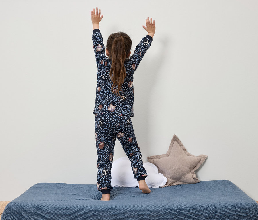Une fille en pyjama bleu marine imprimé se tient sur un lit bleu, les bras levés.