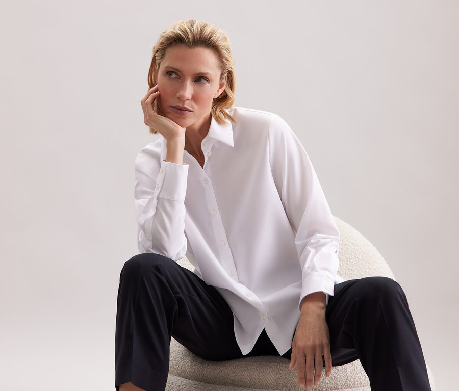 Une femme assise sur une chaise, portant une chemise blanche et un pantalon noir.