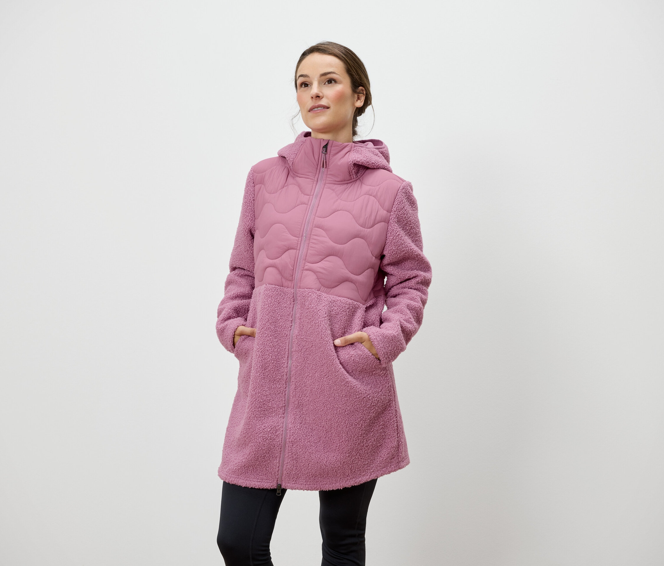 Une femme se tient debout, les mains dans les poches d'un manteau rose à fermeture éclair et à capuche.