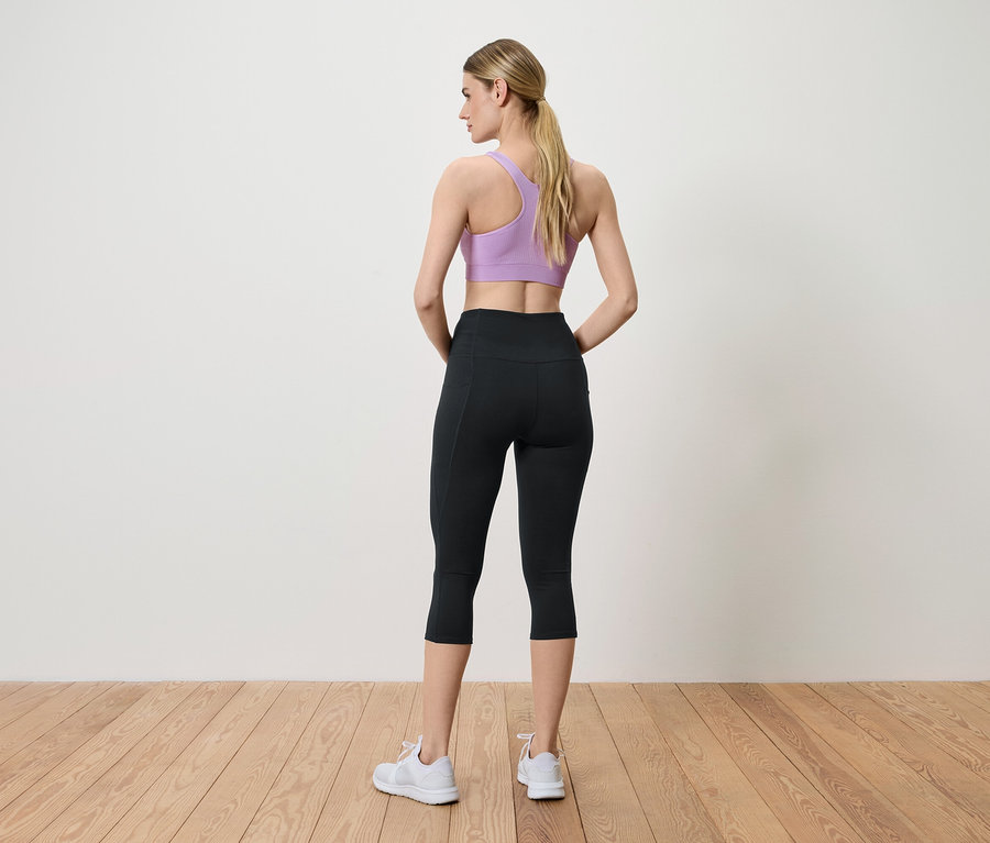 Une femme pose dos à la caméra portant une brassière de sport violette, un legging noir et des baskets blanches.