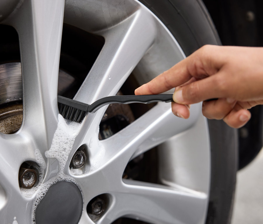 Une main nettoie une jante en aluminium argentée de voiture avec une brosse et de la mousse.
