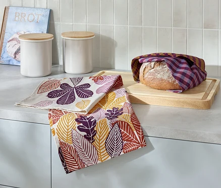 Sur un comptoir de cuisine, une miche de pain enveloppée dans un torchon à carreaux repose sur une planche à découper en bois, à côté de deux torchons avec un imprimé de feuilles, deux boîtes blanches et un livre avec l'inscription Brot.