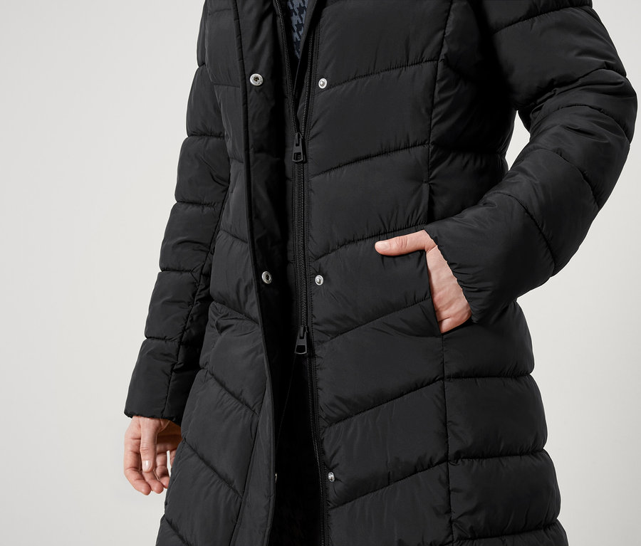 Vue partielle d'une personne portant un manteau noir matelassé avec une fermeture éclair et des boutons. Une main est dans la poche.
