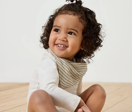 Un bébé souriant est assis sur un plancher en bois et porte un des 5 foulards en mousseline pour bébé.